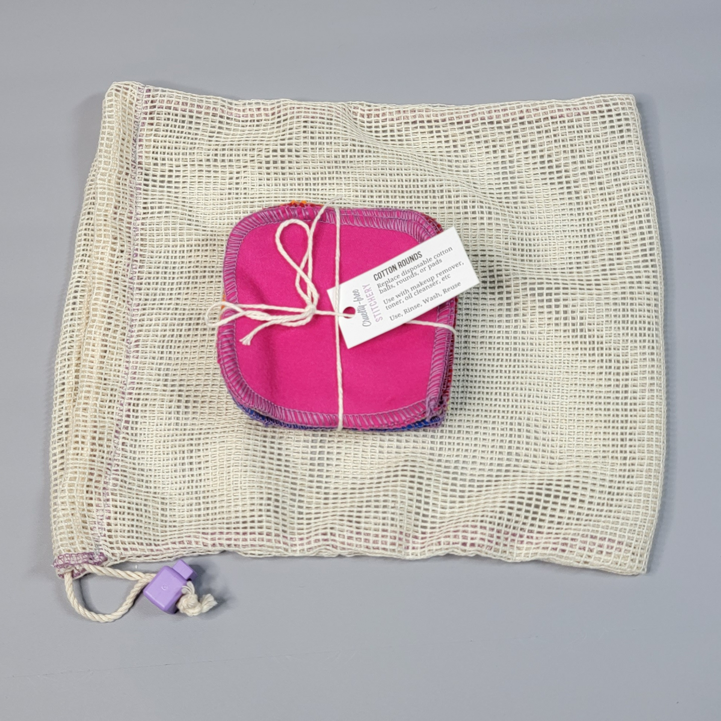 Bundled pack of cotton rounds on top of a mesh drawstring bag. The bag is a natural off-white colored square mesh, with a light purple cord stop.