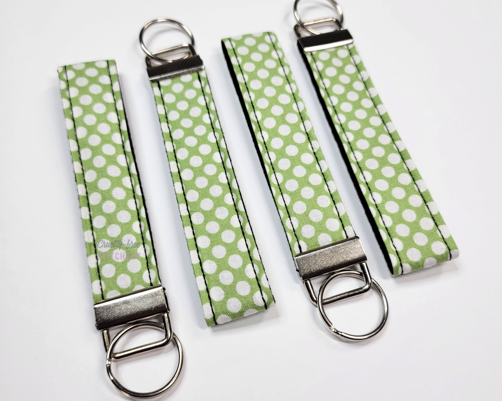 Wristlet key fobs arranged in a row of four with ends alternating. The wristlet is green fabric that has closely-packed white dots printed on. The hardware and key ring are silver.