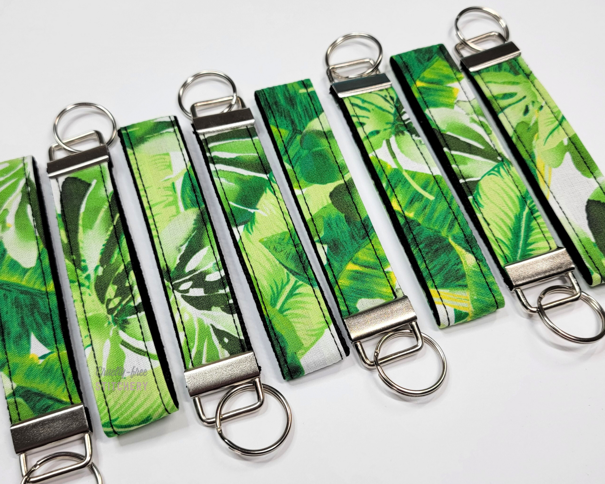 Wristlet key fobs arranged in a row of eight with ends alternating. The wristlet is fabric with large green leaves printed on a barely visible white background. The hardware and key ring are silver.