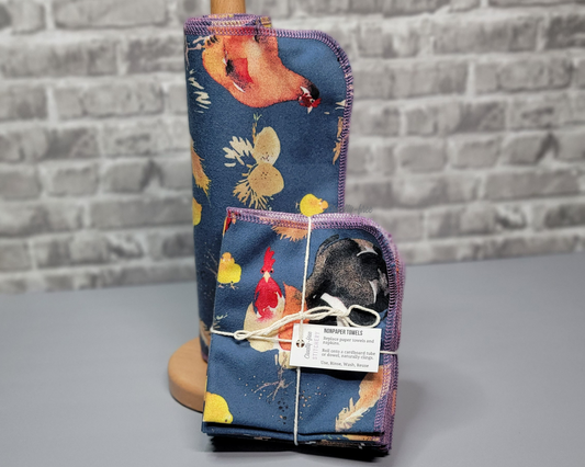 A set of deep blue NonPaper Towels with large chickens, eggs, and little yellow chicks in a realistic painted style. One set is rolled around a wooden holder, the other is folded into a bundle and sitting in front of the holder. 