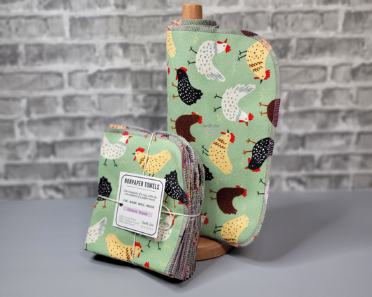 Chickens print NonPaper Towels on a wooden paper towel holder, next to a bundled pack. The print is a light green with white, yellow, brown, and black chickens. Edges are stitched with light purple thread.