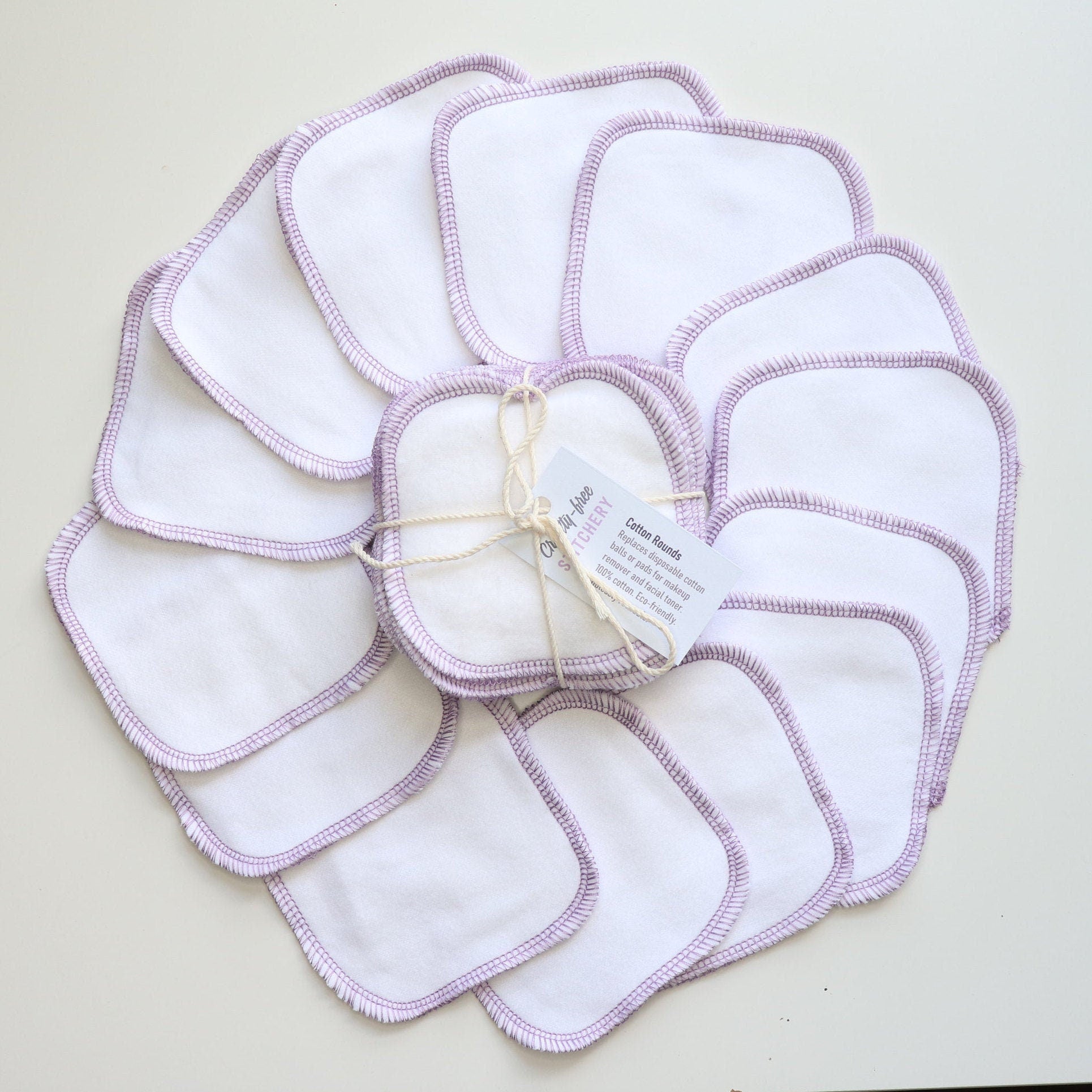 White reusable cotton rounds - arranged in a circle with a bundled pack in the center. They are white fabric that is stitched around the edges in a rounded square shape with light purple thread.