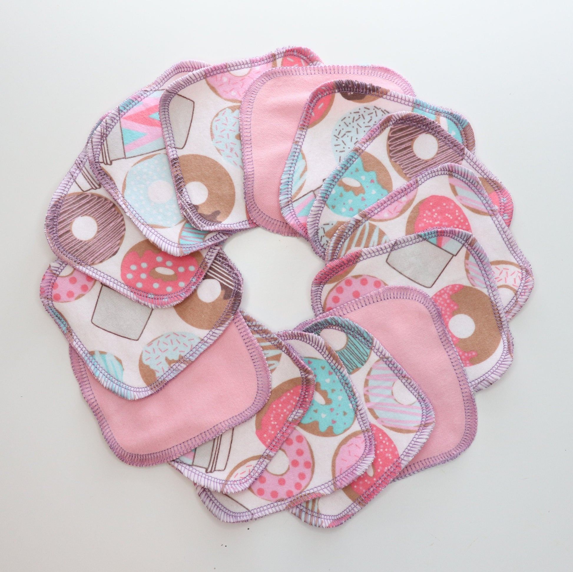 White with donuts and coffee print with light pink on the back - reusable cotton rounds, arranged in a circle. They are a rounded square shape, and stitched together with light purple thread.