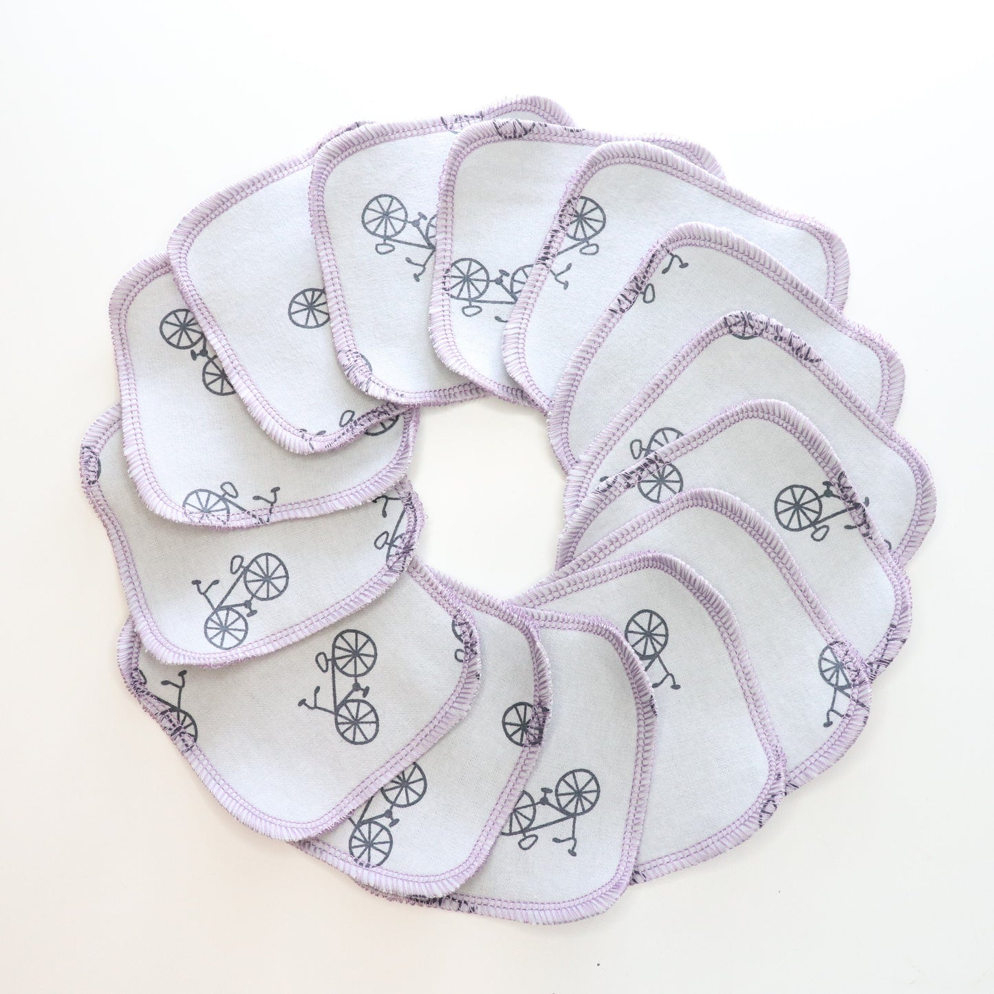 Reusable cotton rounds arranged in a circle. This print is a light grey with scattered black bicycles. Stitched together with light purple thread in a rounded square shape.