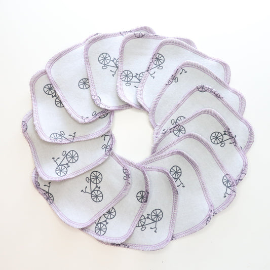 Reusable cotton rounds arranged in a circle. This print is a light grey with scattered black bicycles. Stitched together with light purple thread in a rounded square shape.