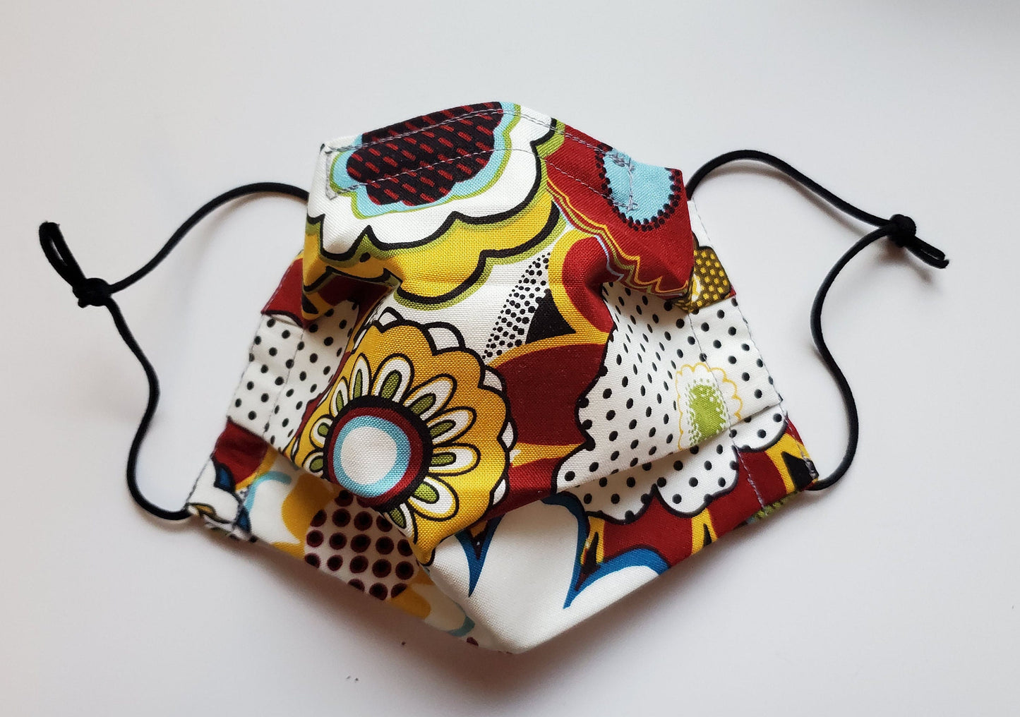 A cloth face mask in a bold 70s print with various outlined flowers and polka dots in reds, yellows, light blue, light green, and black and white. The mask is pleated with black elastic ear loops.