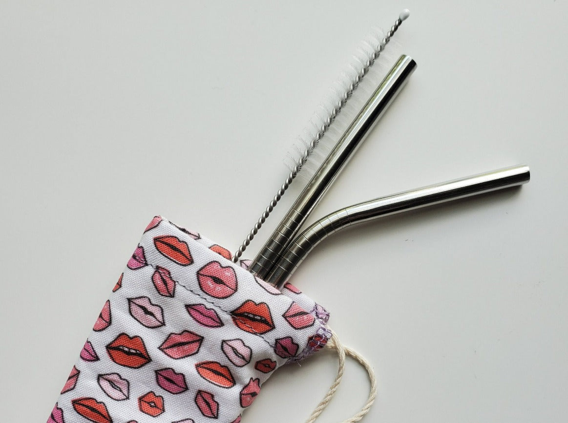 Reusable straw pouch with stainless steel straws sticking out the top. The fabric of the pouch is white with tiny cartoon lips in varying shades of pink, and it has a drawstring closure.