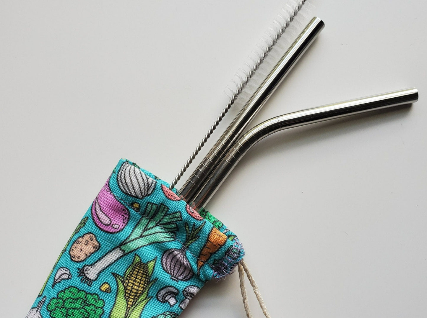 Reusable straw pouch with stainless steel straws sticking out the top. The fabric of the pouch is teal blue with various cartoon brightly colored vegetables like broccoli, carrots, radishes, etc and it has a drawstring closure.
