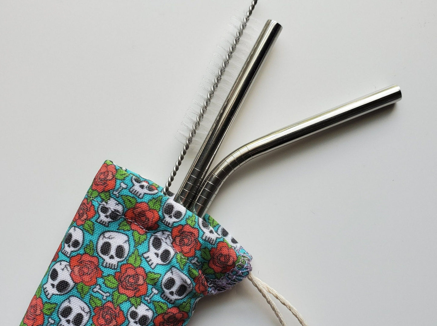 Reusable straw pouch with stainless steel straws sticking out the top. The fabric of the pouch is blue with tiny white skulls and red roses, and it has a drawstring closure.