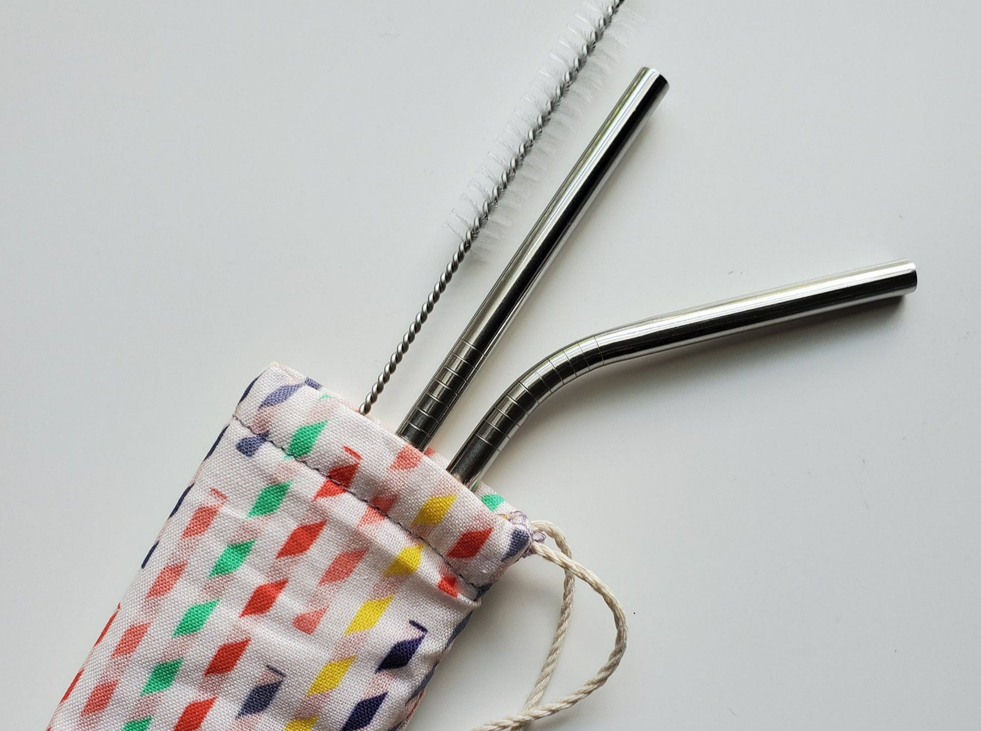 Reusable straw pouch with stainless steel straws sticking out the top. The fabric of the pouch is white with multicolored paper straws printed on, and it has a drawstring closure.