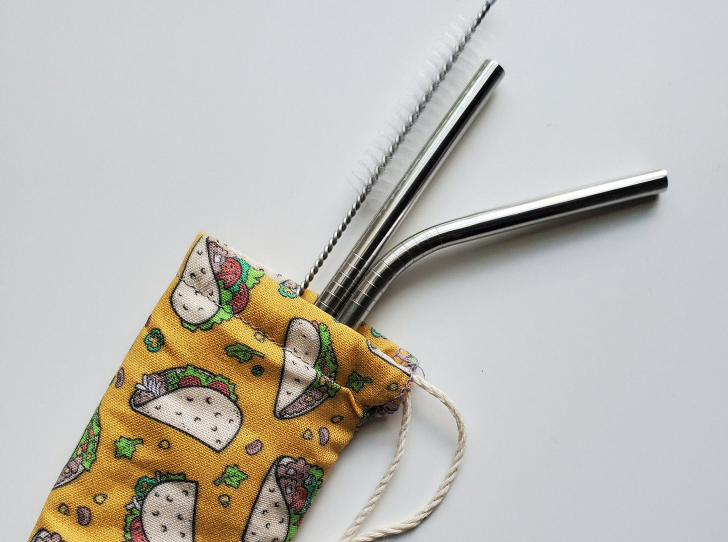 Reusable straw pouch with stainless steel straws sticking out the top. The fabric of the pouch is mustard yellow with small cartoon tacos and scattered toppings, and it has a drawstring closure.