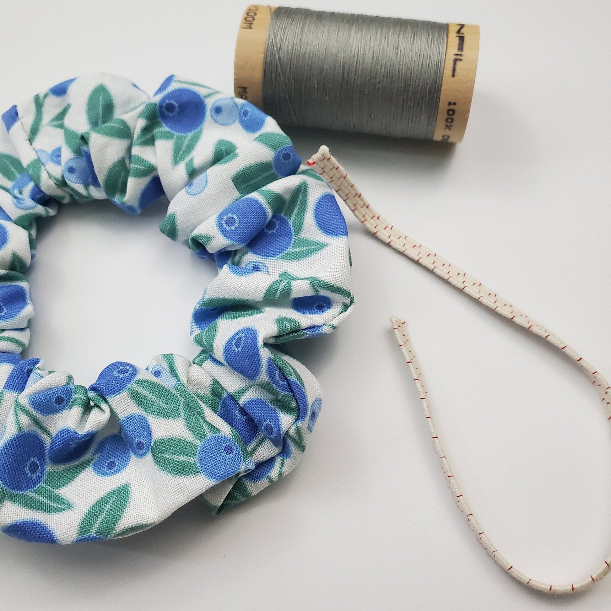 Blueberry print Scrunchie shown next to a wooden spool of grey thread, and a piece of the biodegradable elastic.