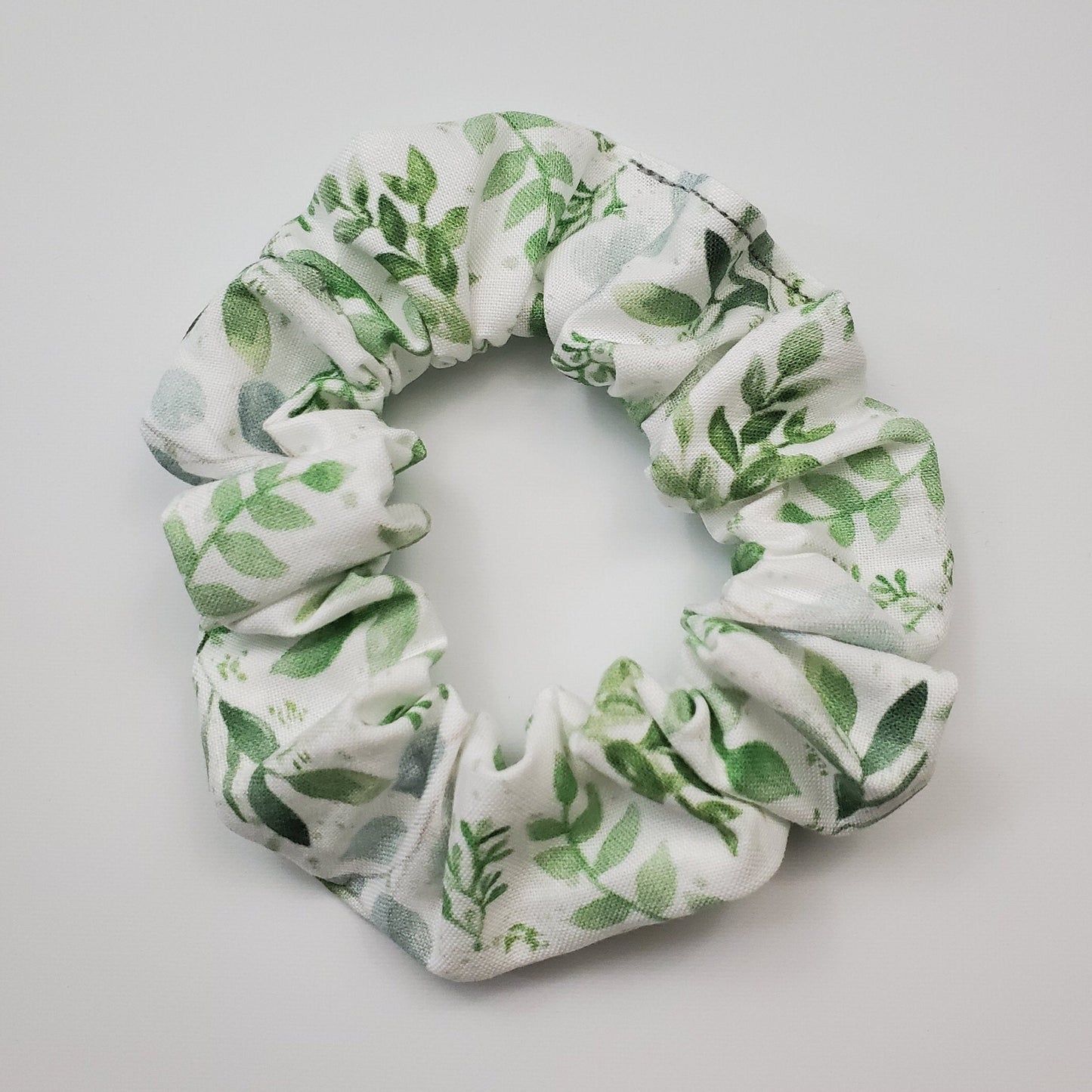 White scrunchie with light green eucalyptus leaves. The leaves appear to be painted in watercolor and have various vines and other leaves around.
