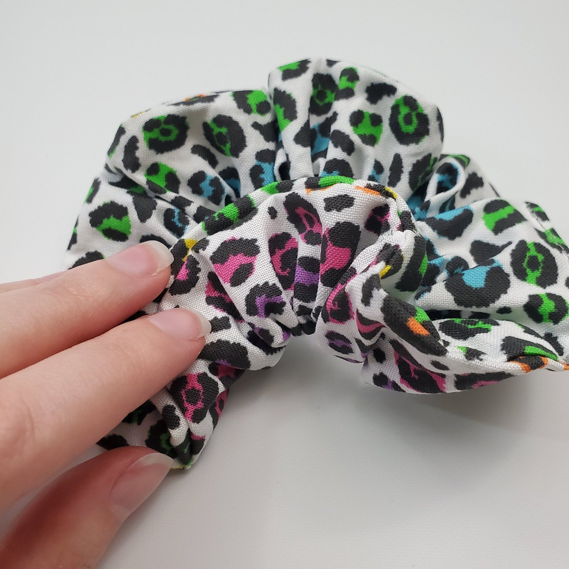 The rainbow cheetah print scrunchie being held to show both sides. This one has more green and blue on one side and pink and purple on the other.
