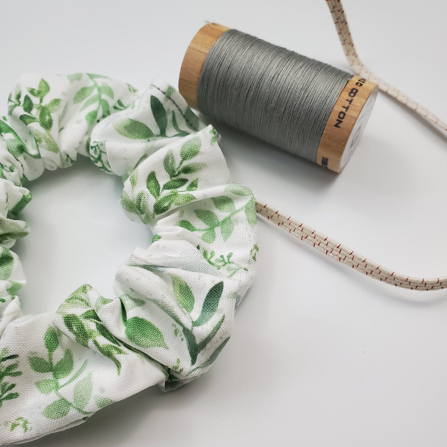 Scrunchie shown next to a wooden spool of grey thread, and a piece of the biodegradable elastic.