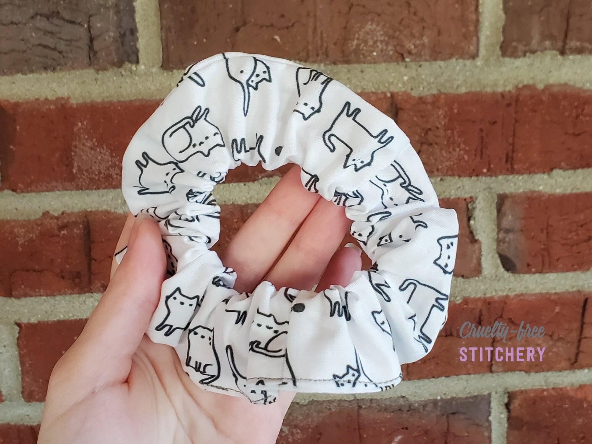 White scrunchie with black outlined cartoon cats in various poses. Sitting, lying down, playing with yarn, showing butt, etc.