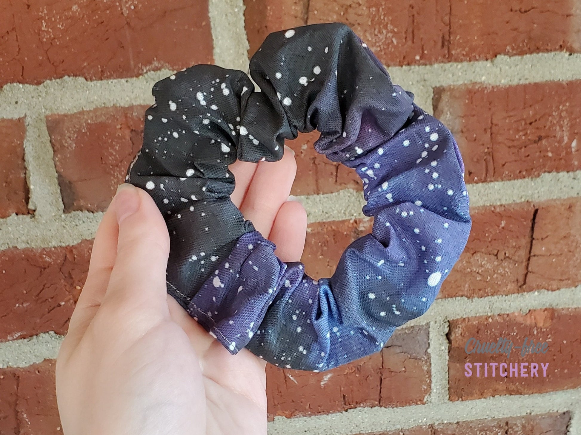 A galaxy print scrunchie. The background color is various shades of black, dark blue, and dark purple with small white dots.