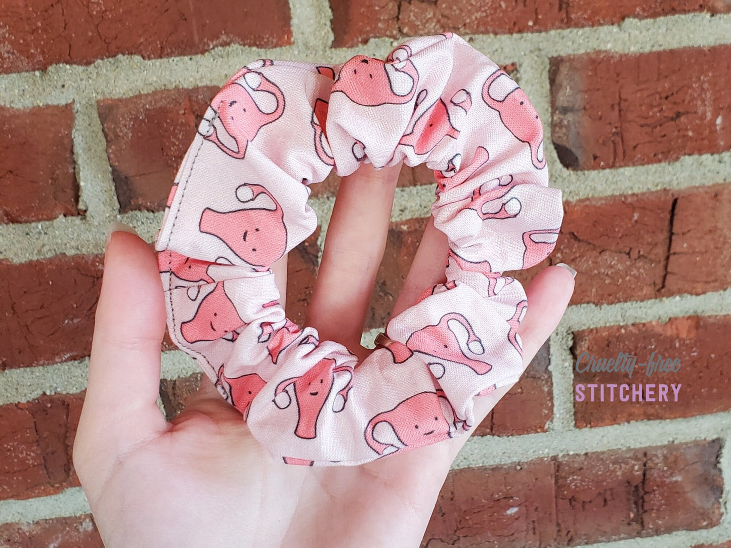 A light pink scrunchie with darker pink smiling uteruses. Held in my hand on a red brick background.