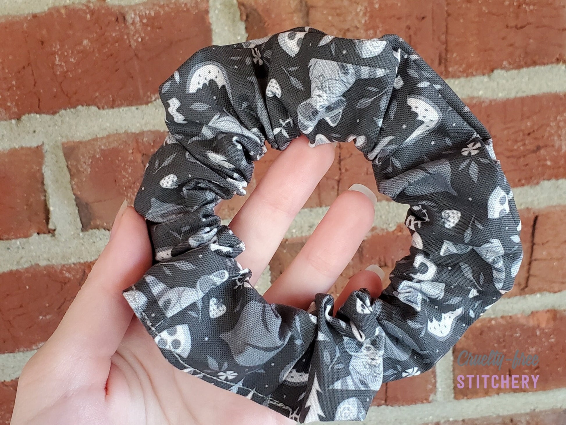 Black and grey raccoons print scrunchie. Features cute raccoons, trash bags, and scattered food. Pictured in my hand on a red brick background.