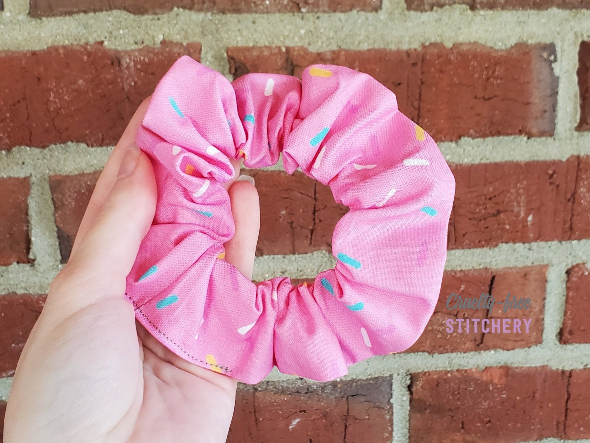 A bright pink scrunchie with scattered sprinkles in blue, white, yellow, and light pink. Pictured in my hand on a red brick background.