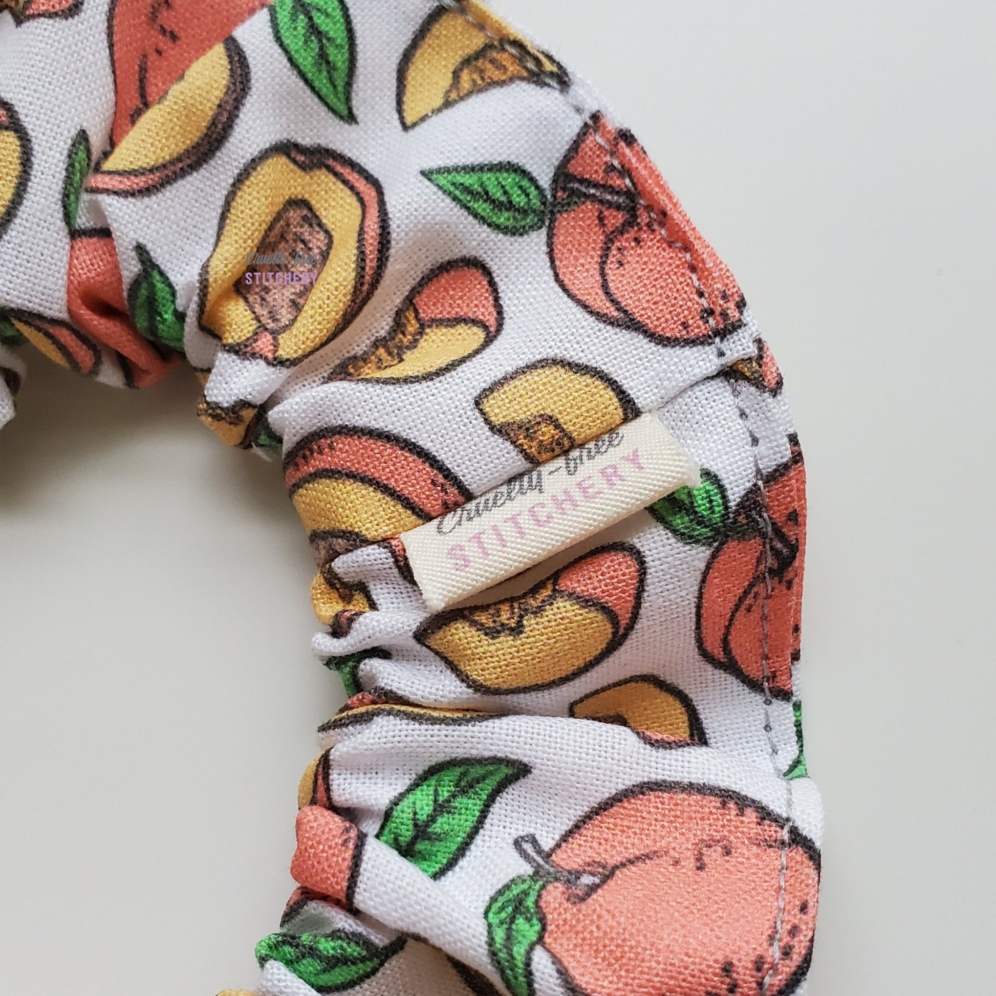 Close-up of the peaches print scrunchie with a small white tag with the Cruelty-Free Stitchery logo.
