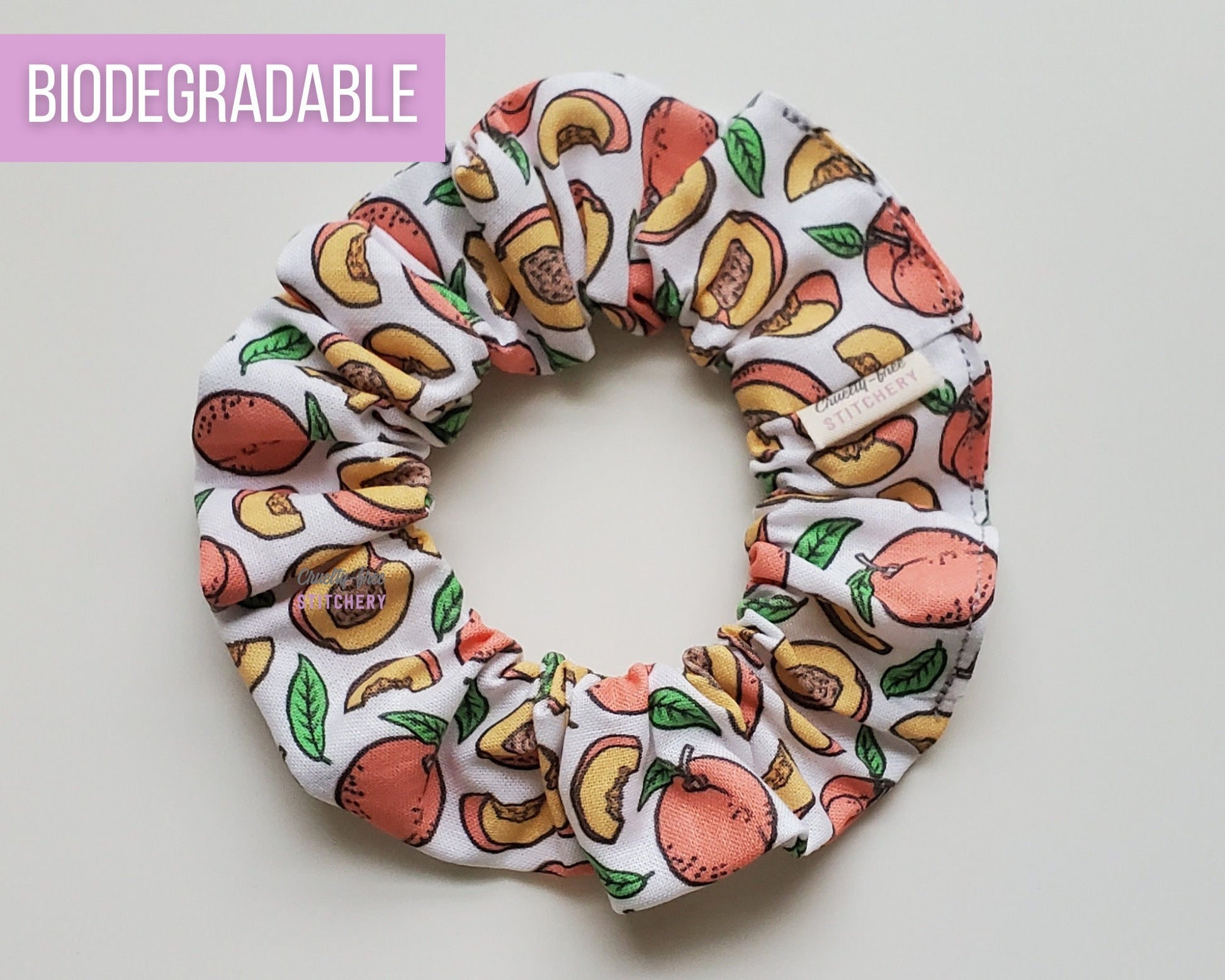 A scrunchie with cartoonish peaches printed on a white fabric. Some whole peaches, some slices, some pits, and leaves. 