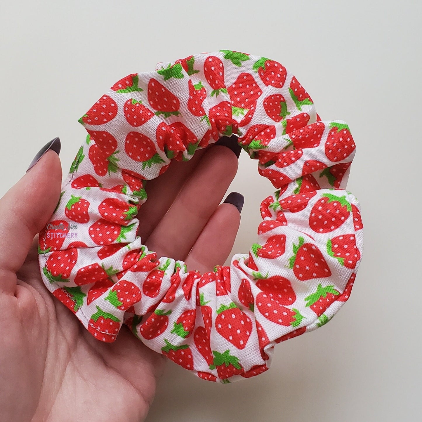 Strawberry print scrunchie in my hand to show the size. The fabric part is the width of two fingers, and the entire scrunchie is as wide as my fingers and half of my palm.