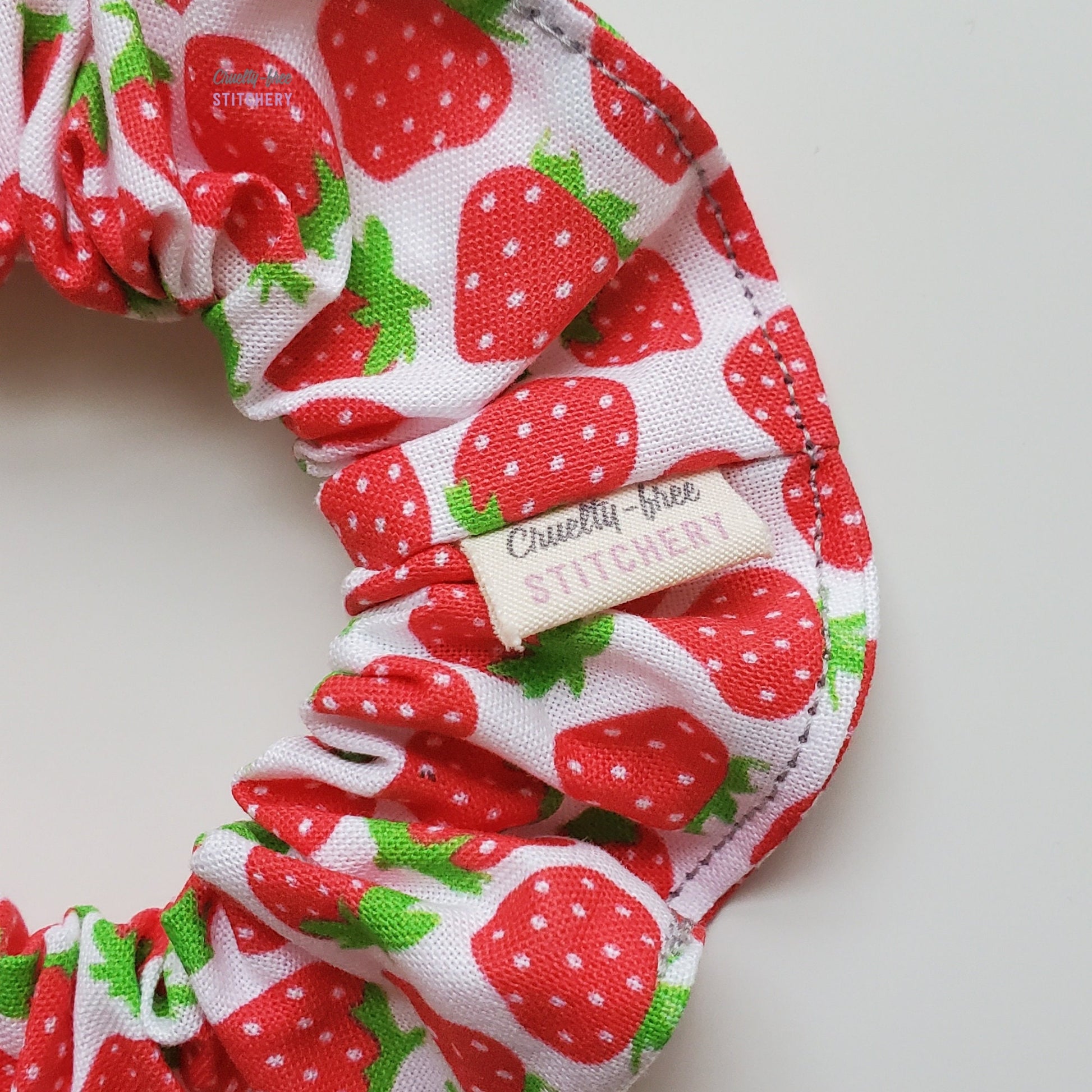 Close-up image of the side of the strawberry print scrunchie, with a small tag in the seam that has the Cruelty-Free Stitchery logo.