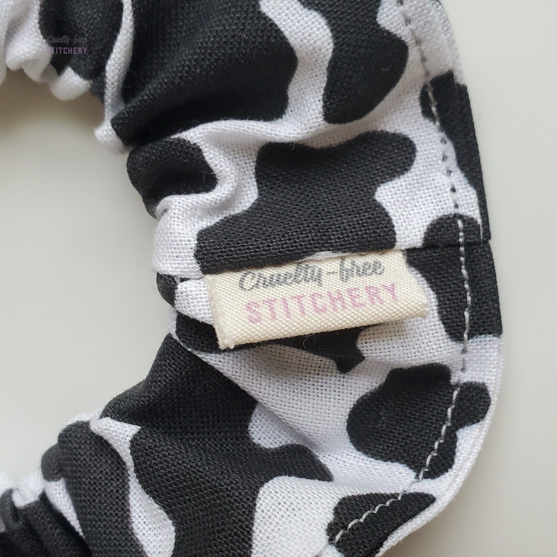 Close-up of the cow print scrunchie with a small white tag with the Cruelty-Free Stitchery logo.