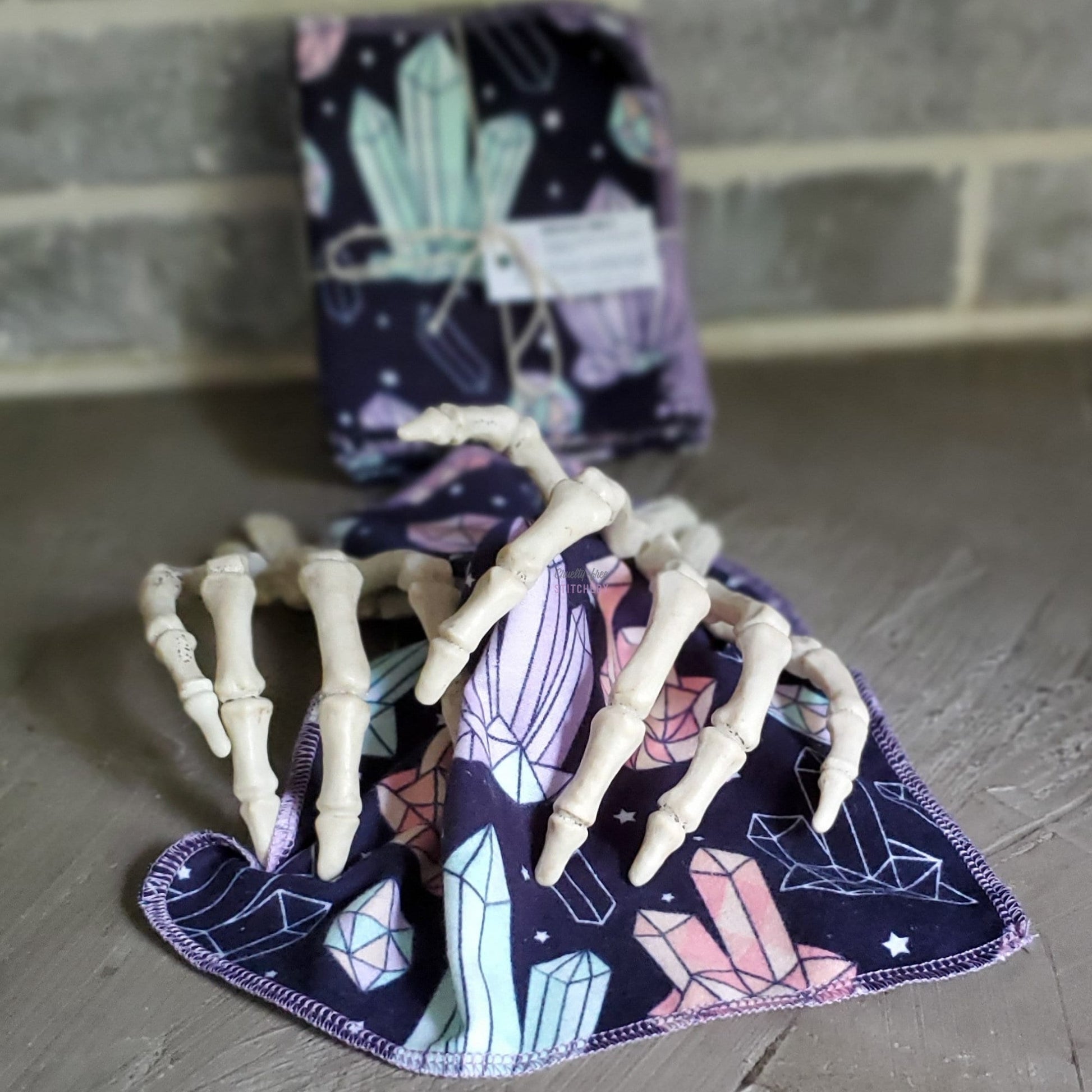 A crystals print NonPaper Towel with two fake skeleton hands as if they are drying off with it. A bundled pack of them is blurred in the background.