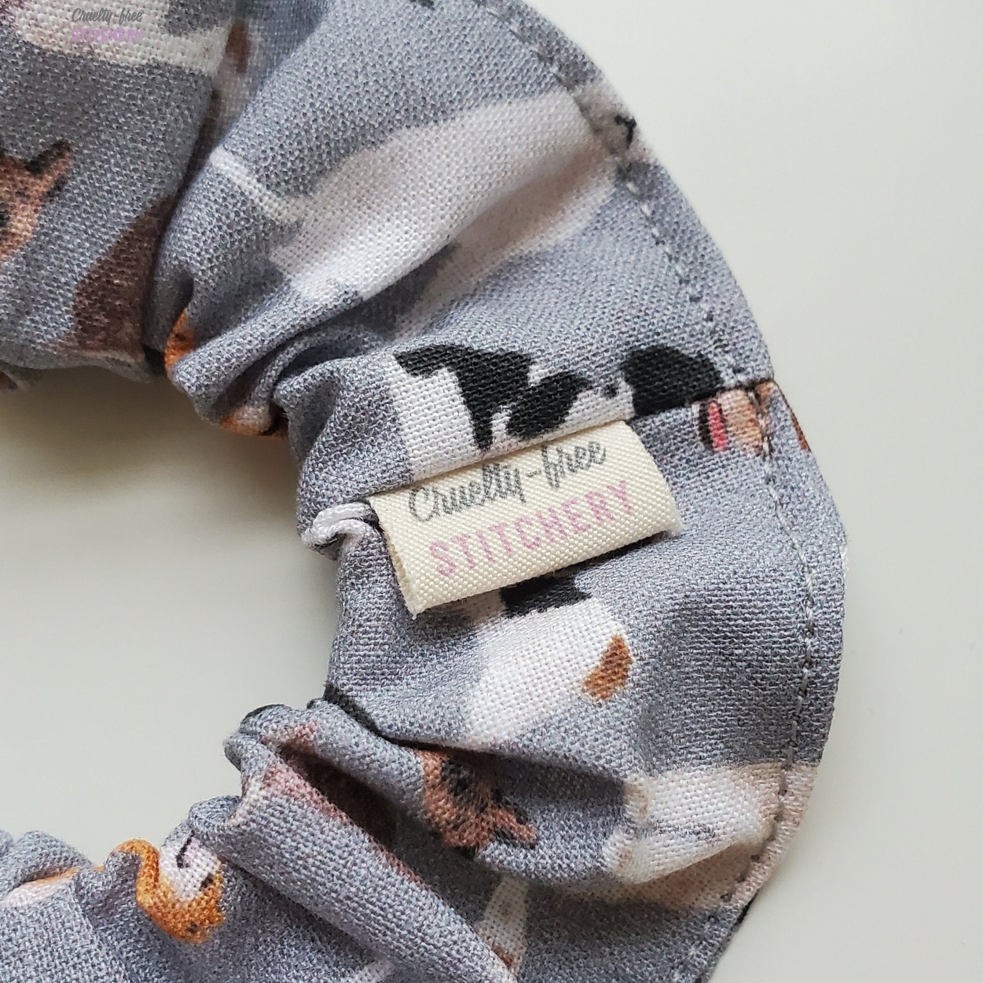 Close-up of the grey with dogs print scrunchie with a small white tag with the Cruelty-Free Stitchery logo.