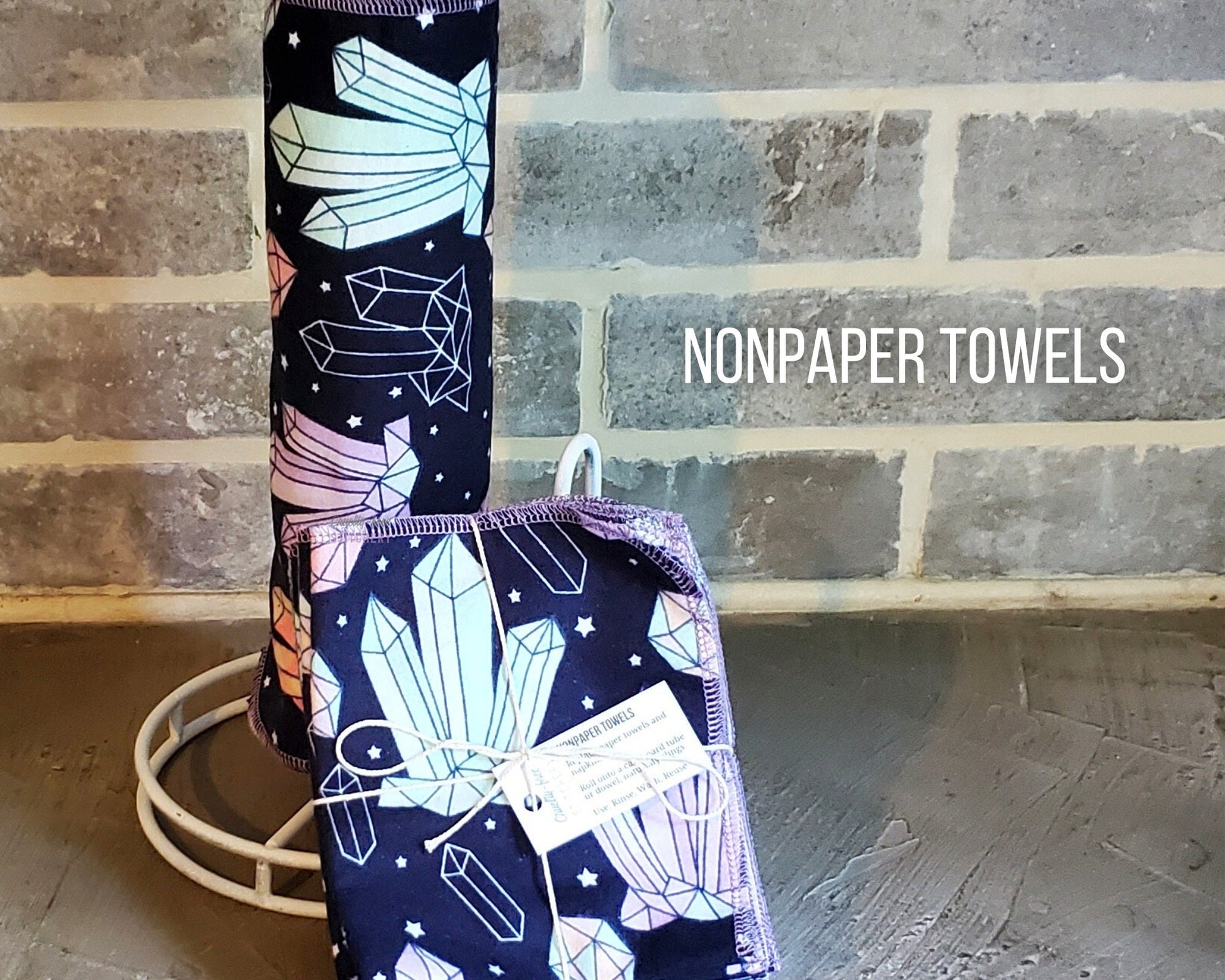 Crystals Print NonPaper Towels. They are rolled onto a paper towel holder next to a pack of folded ones. The print is sketched crystals in light blue, pink, and lavender with tiny stars on a navy blue background.