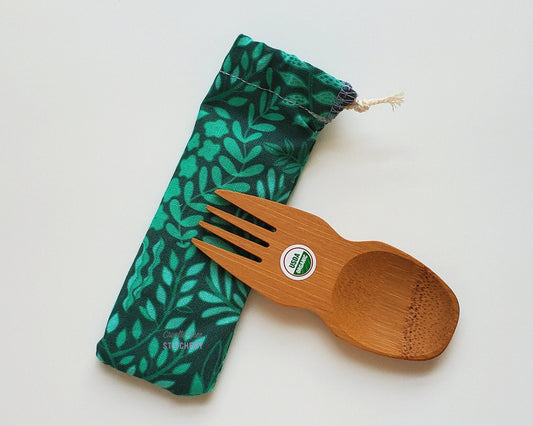 Reusable bamboo spork with pouch. The pouch is dark green with lighter green leaves and vines. The pouch is sitting diagonally with the spork partially on top pointing the other way. The spork is small, a double ended fork and spoon.