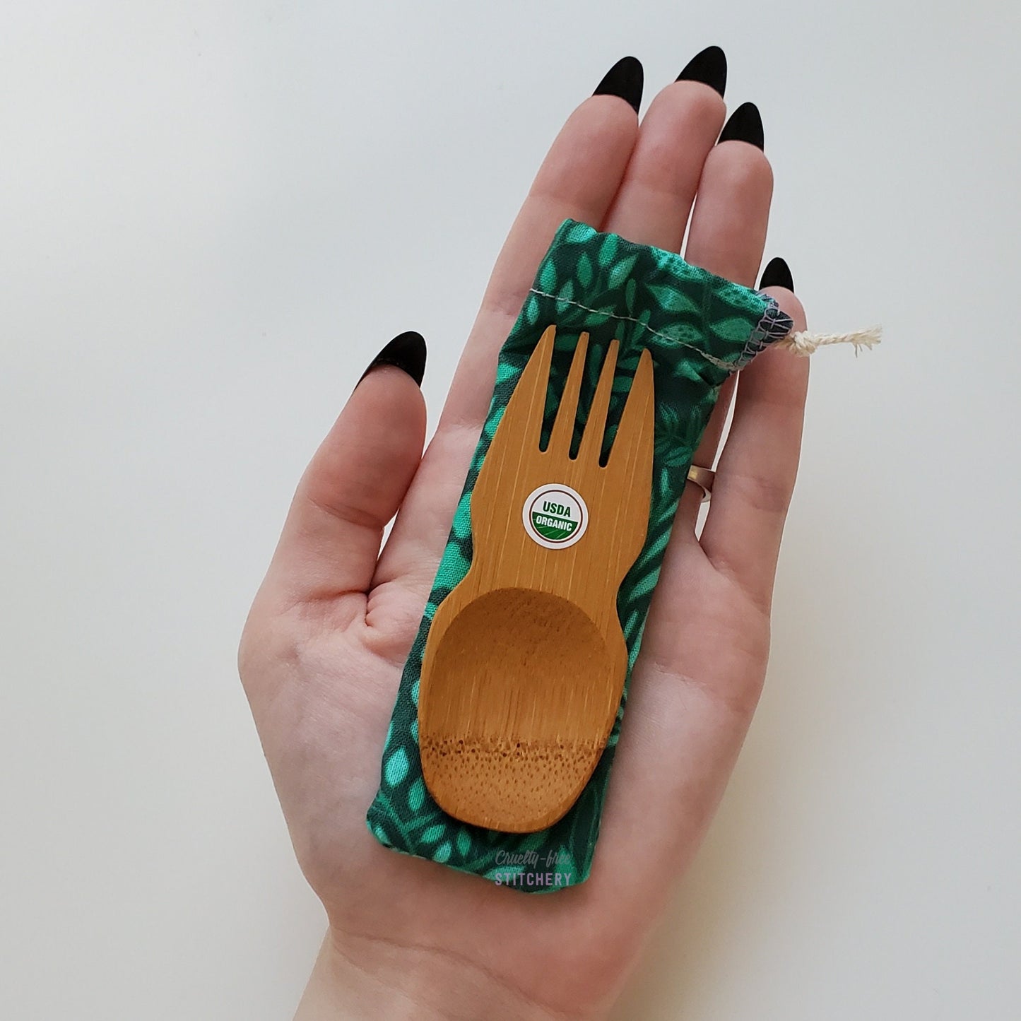 Reusable bamboo spork with pouch. Green with leaves print fabric pouch with bamboo spork on top, laid on a hand for size reference. The spork is slightly longer than the fingers.