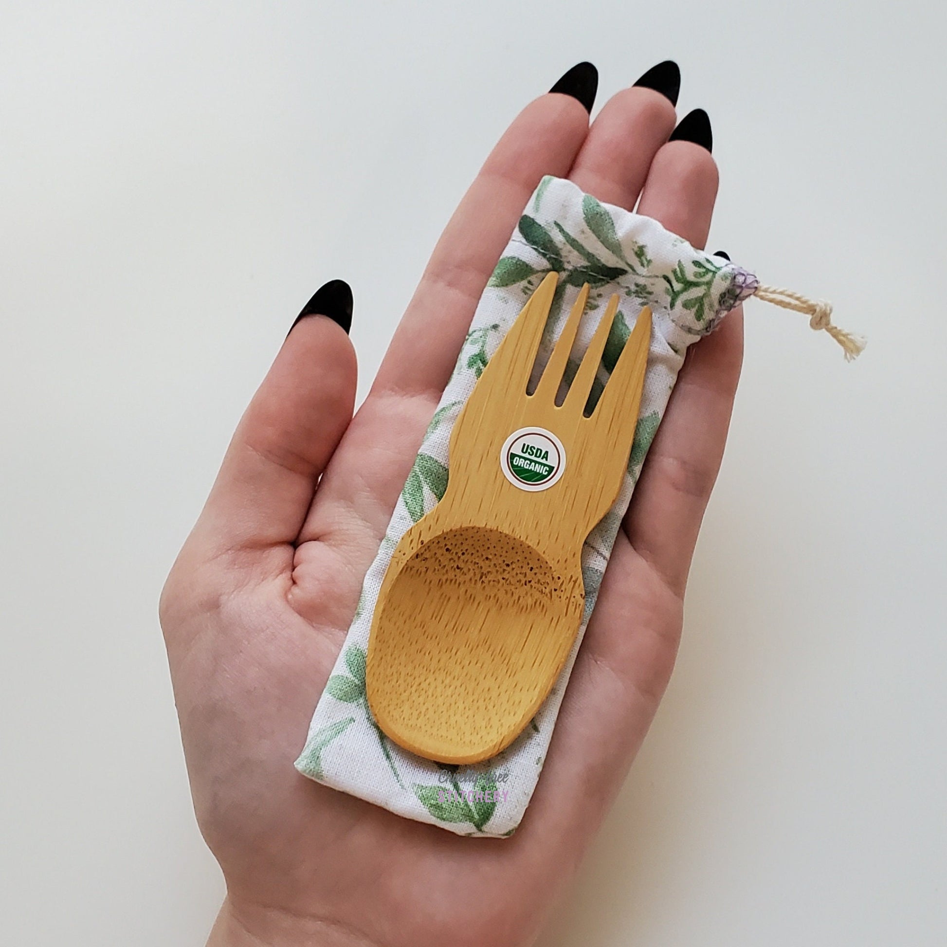 Reusable bamboo spork with pouch. Eucalyptus print fabric pouch with bamboo spork on top, laid on a hand for size reference. The spork is slightly longer than the fingers.