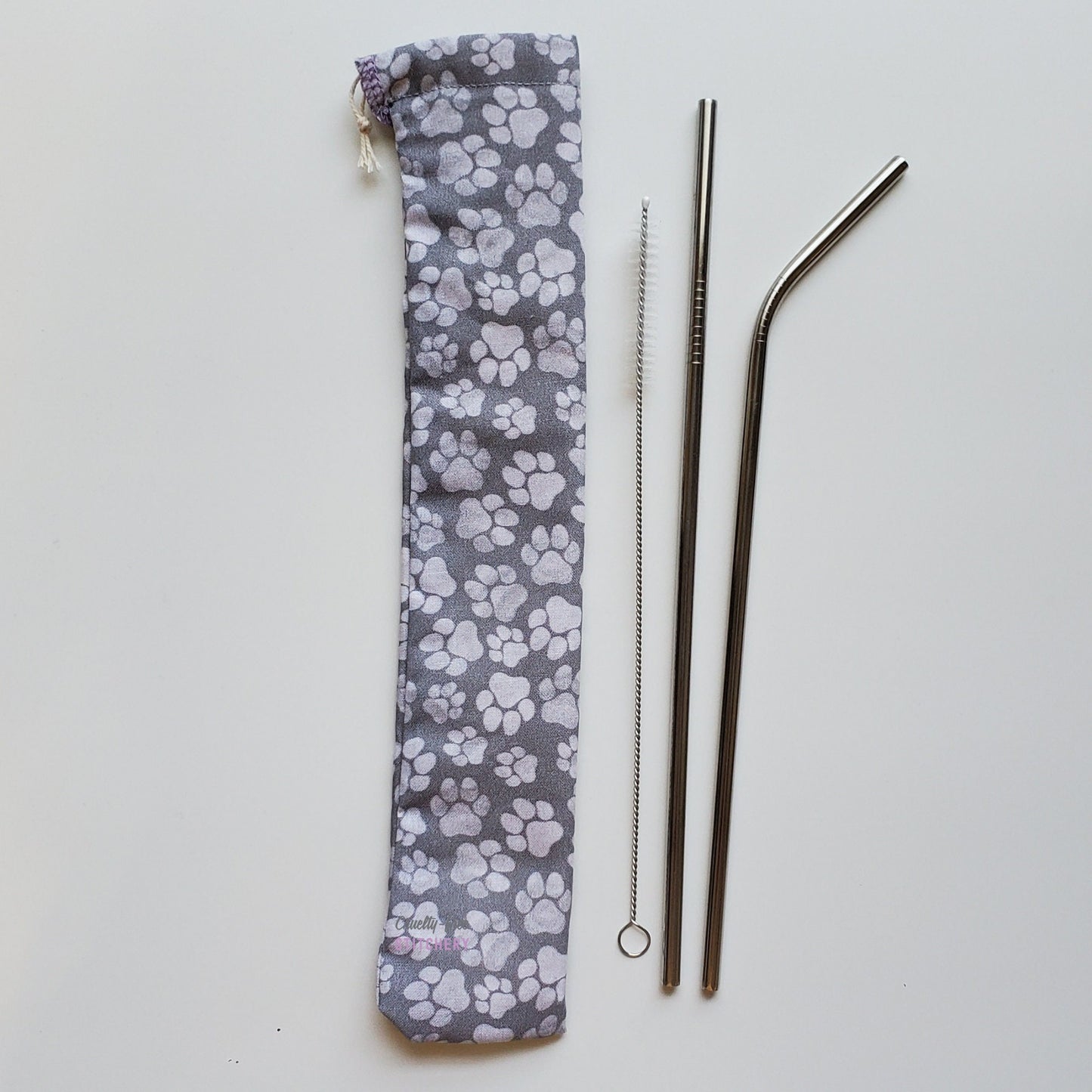 Reusable straw pouch in the same paw print laying vertically next to a straw cleaner brush, a straight stainless steel straw, and a bent stainless steel straw.