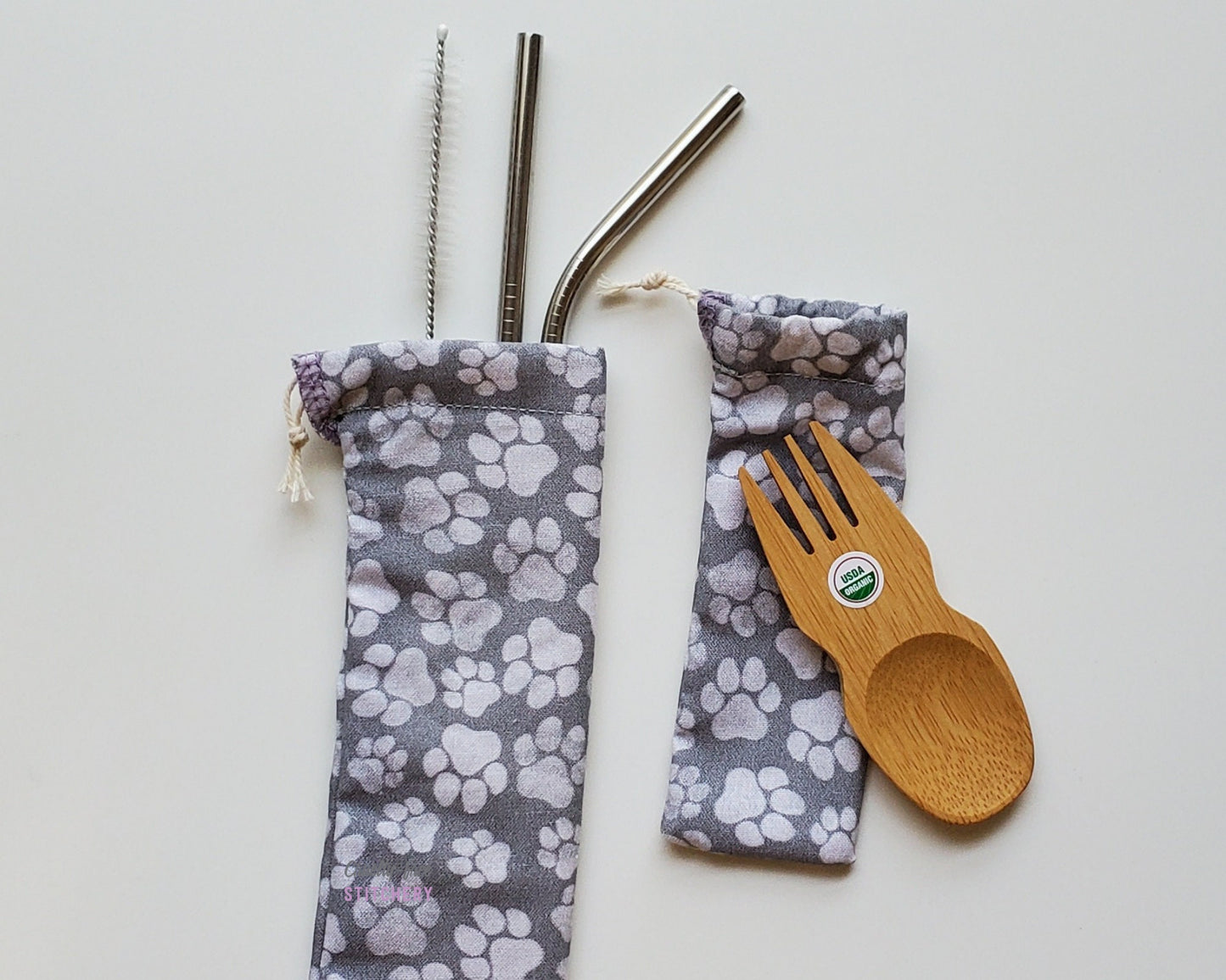 Reusable bamboo spork and stainless steel straw pouch set. The pouches are both grey with lighter grey paw prints scattered in various sizes.