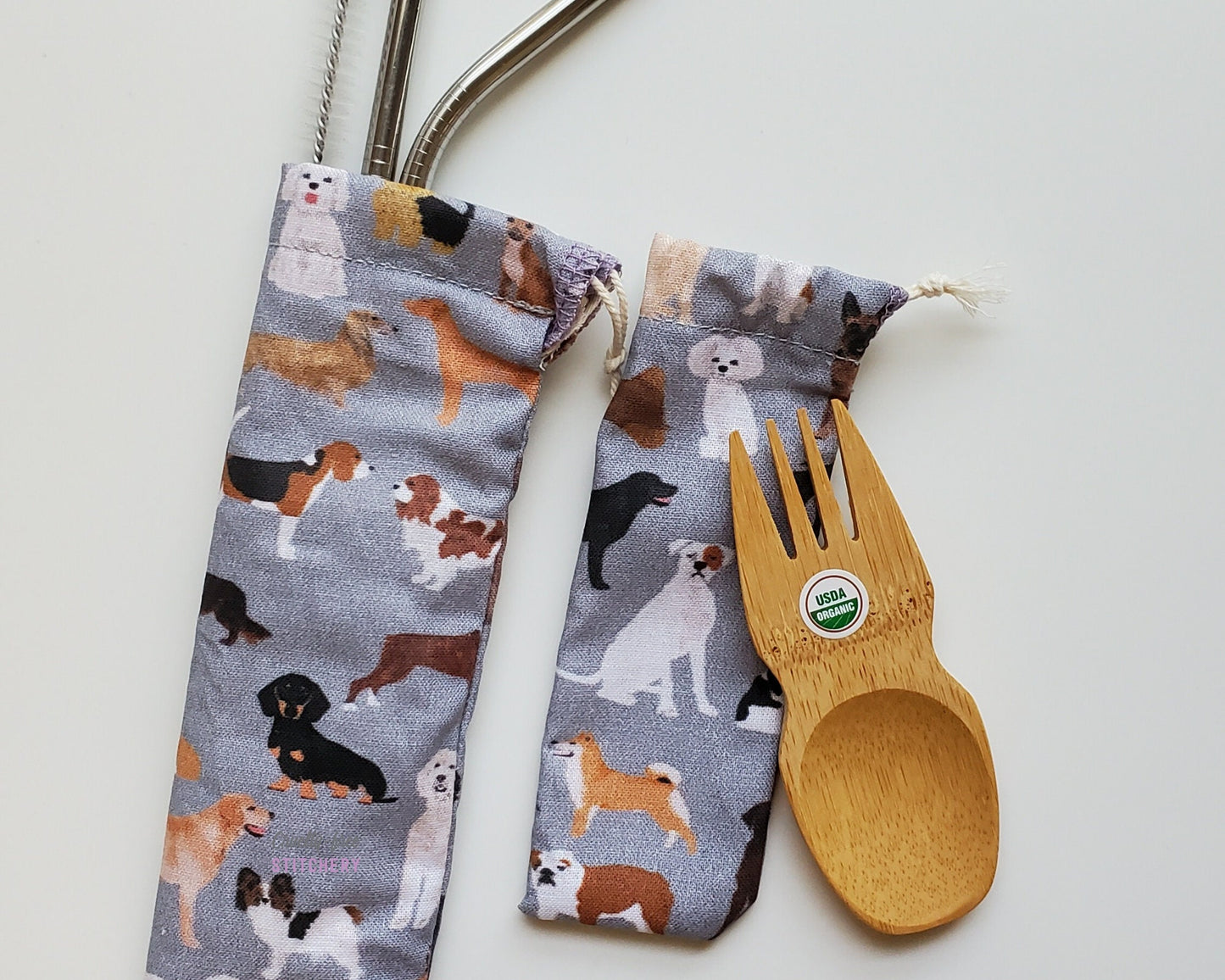 Reusable bamboo spork and straw pouch set. The pouches are a grey with assorted dogs printed on the fabric, such as beagles, basset hounds, dachshunds, pomeranians, golden retrievers, and more.