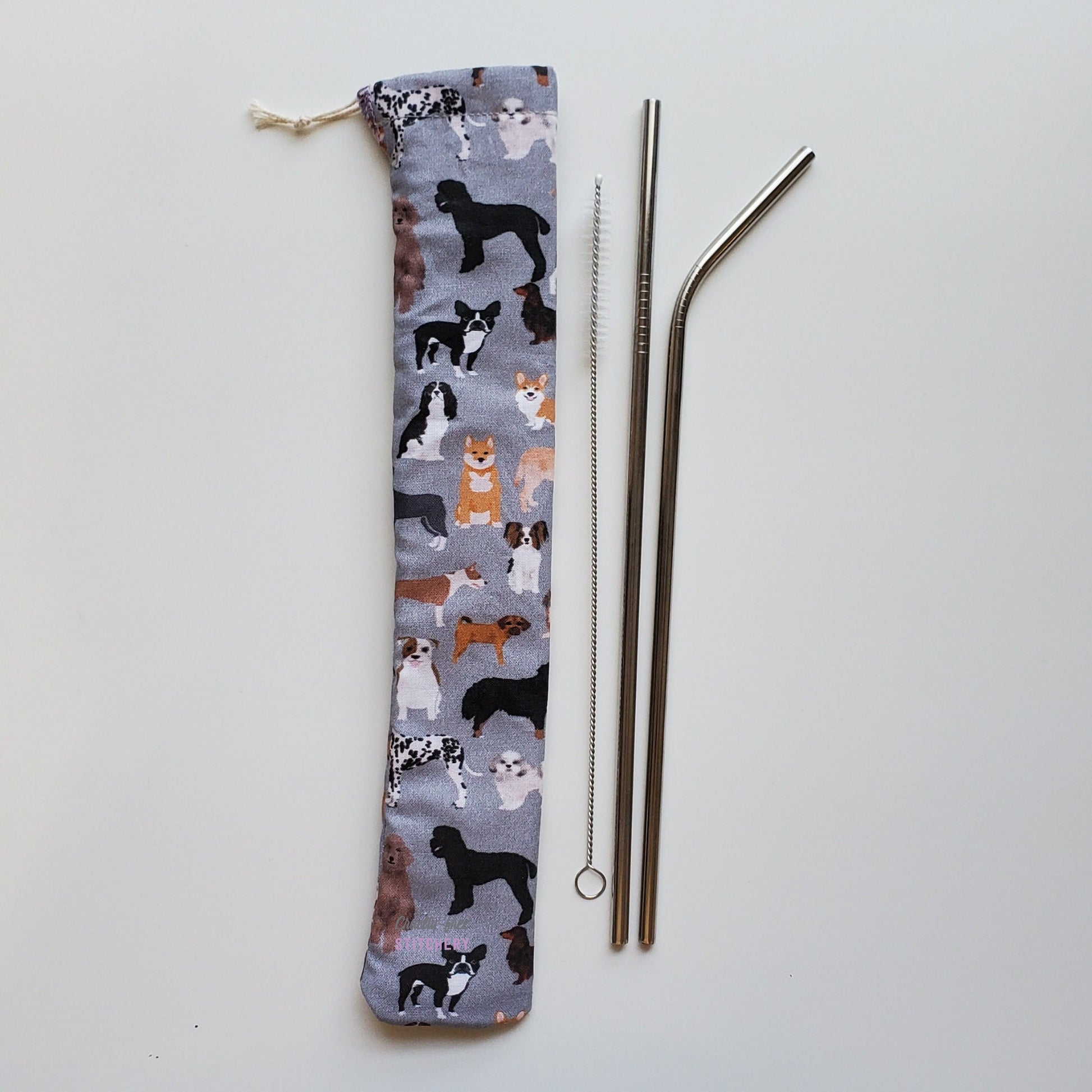 Reusable straw pouch in the same dog print laying vertically next to a straw cleaner brush, a straight stainless steel straw, and a bent stainless steel straw.