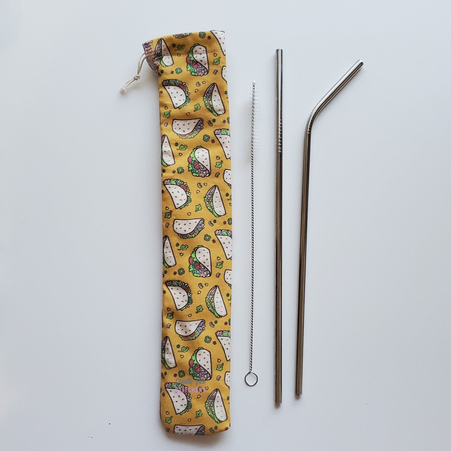 Reusable straw pouch in the same taco print laying vertically next to a straw cleaner brush, a straight stainless steel straw, and a bent stainless steel straw.