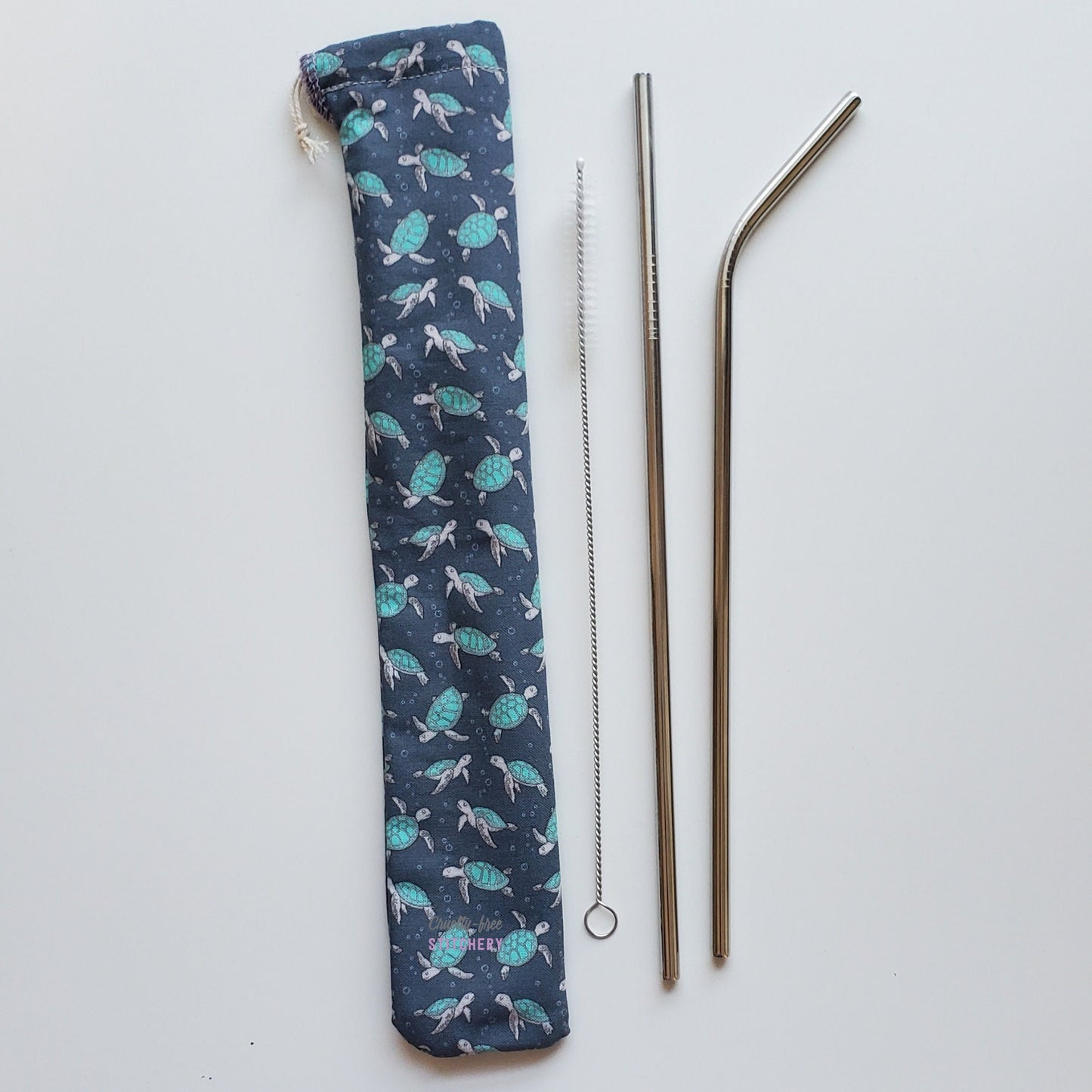Reusable straw pouch in the same turtle print laying vertically next to a straw cleaner brush, a straight stainless steel straw, and a bent stainless steel straw.