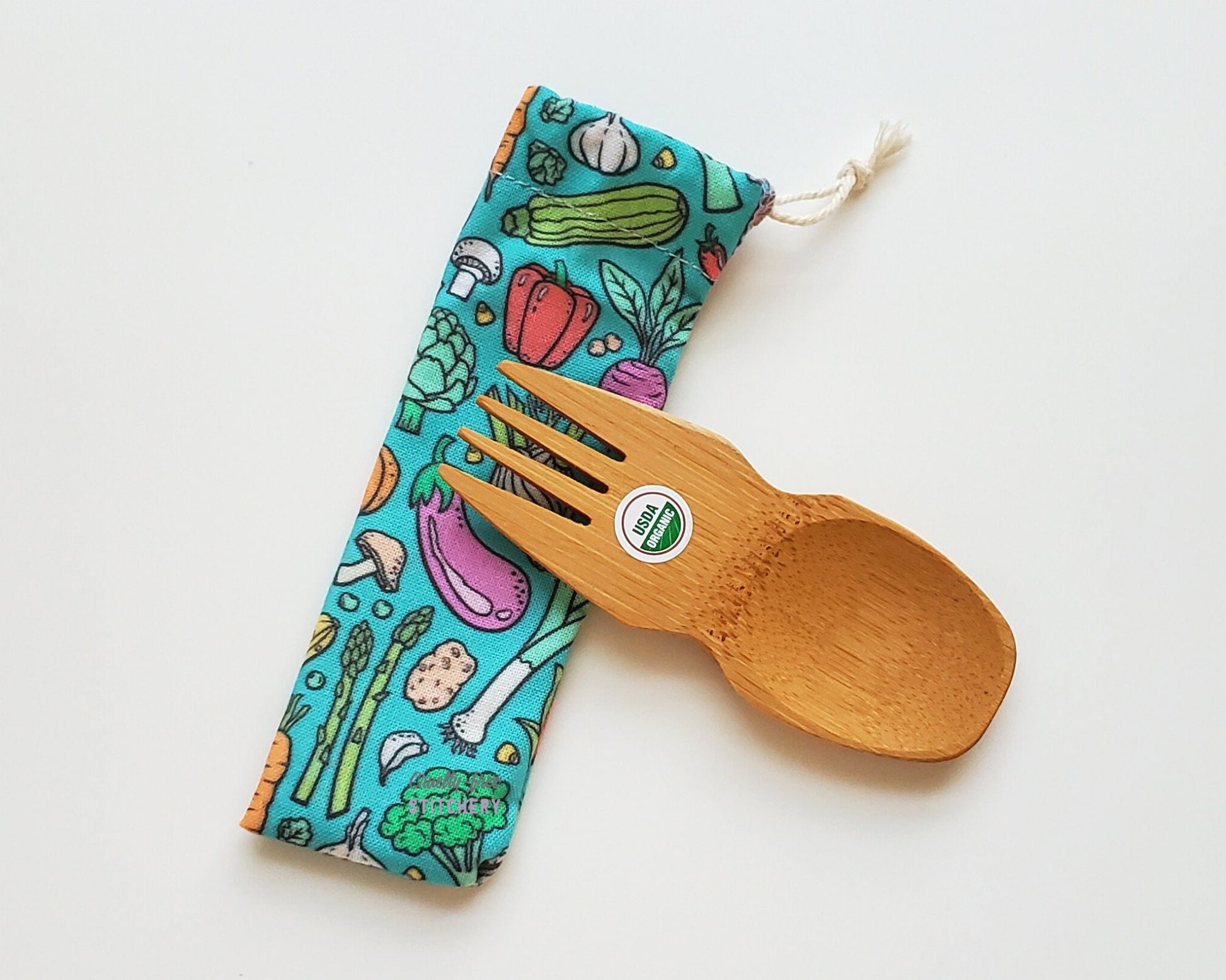 Veggie print reusable spork pouch. The pouch is sitting diagonally with the spork partially on top pointing the other way. The fork end of the spork is pointing to the left.