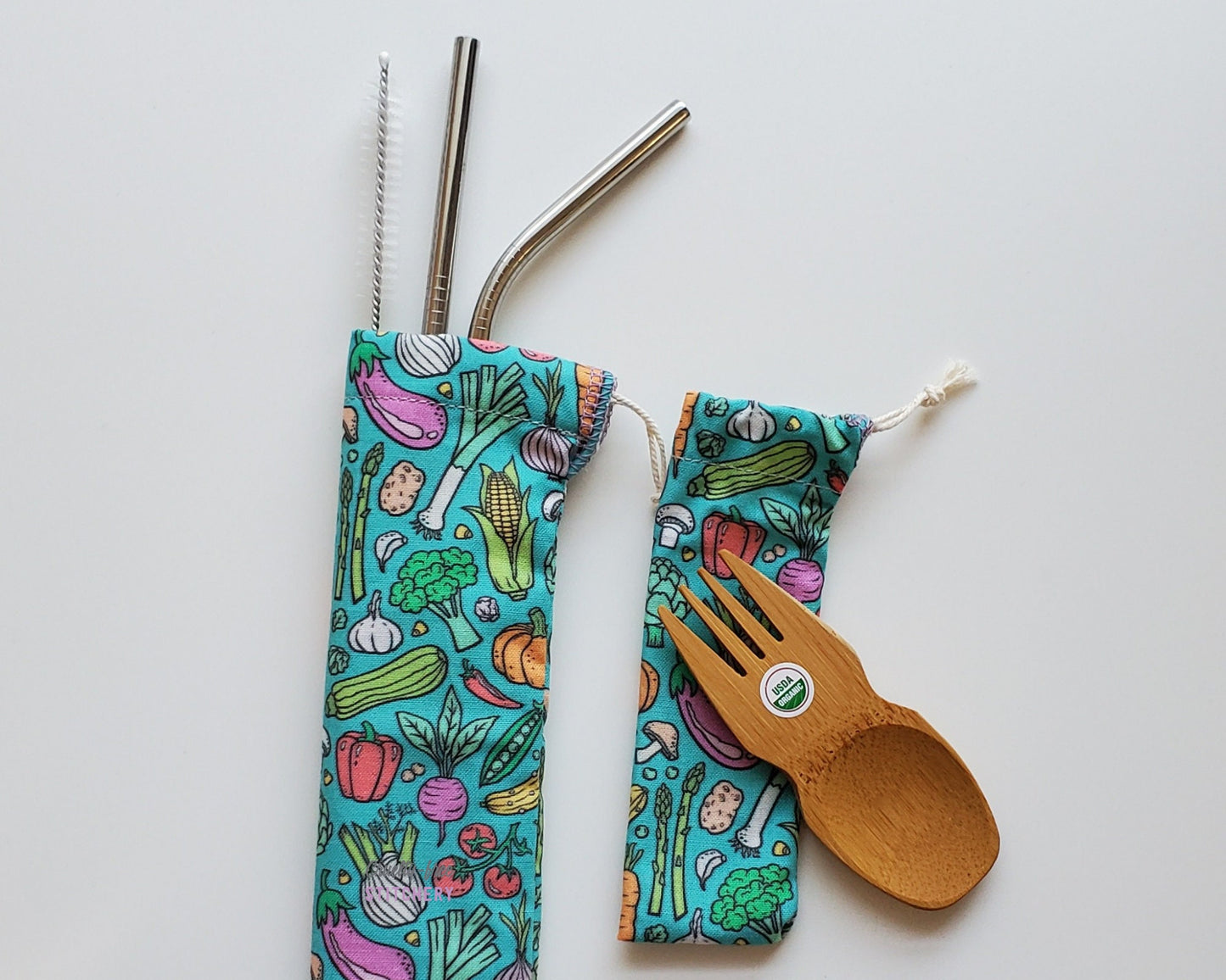 Reusable bamboo spork and stainless steel straw pouch set. The pouches are both a teal blue background with cartoon vegetables printed on the fabric. Eggplants, radishes, peppers, tomatoes, asparagus, broccoli, mushrooms, and more.