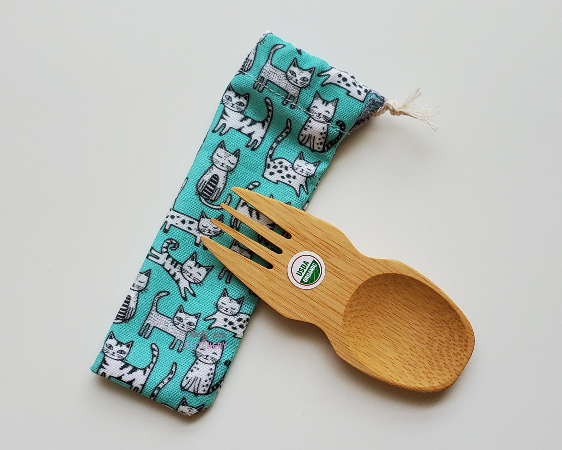 Blue with white cats print reusable spork pouch. The pouch is sitting diagonally with the spork partially on top pointing the other way. The fork end of the spork is pointing to the left.