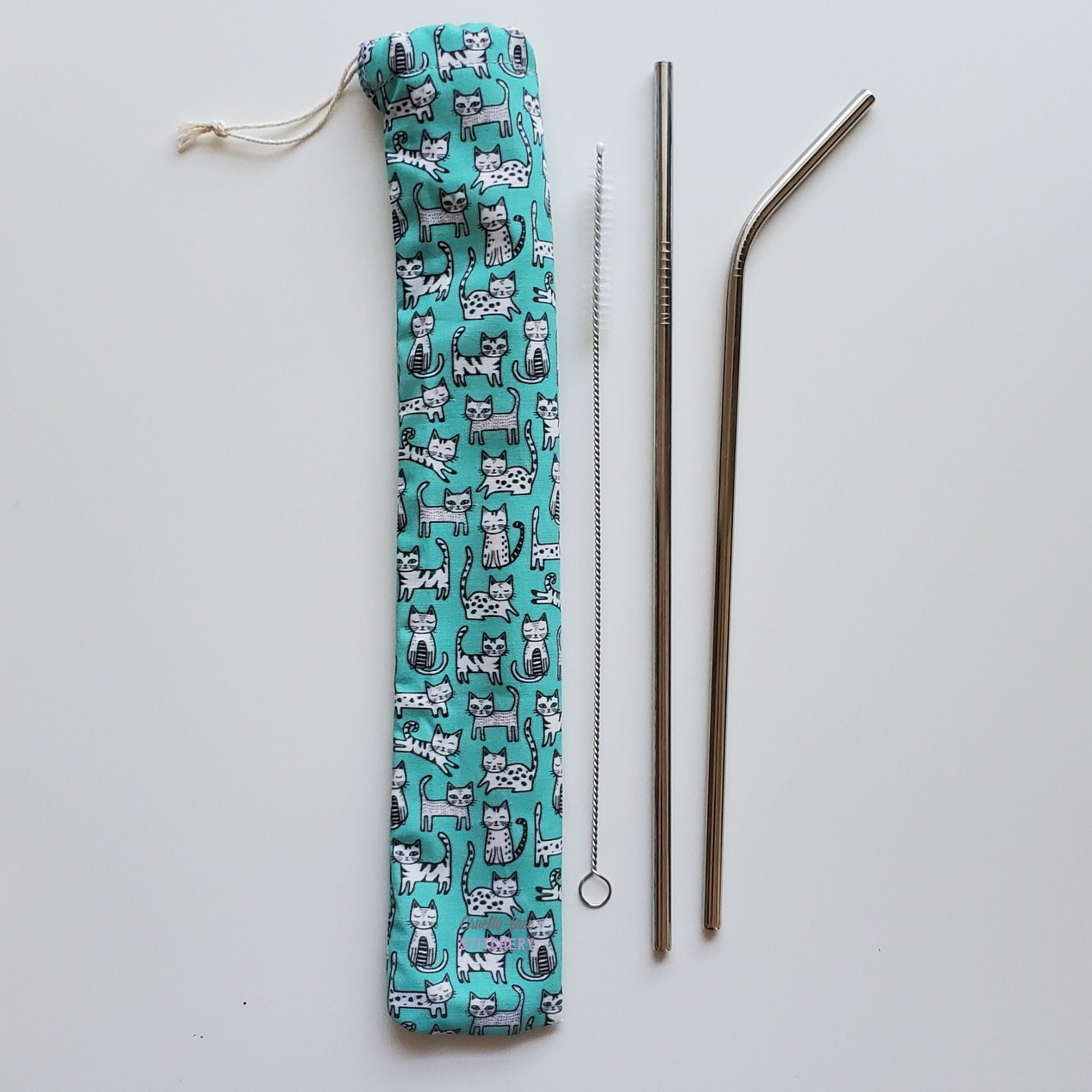 Reusable straw pouch in the same cat print laying vertically next to a straw cleaner brush, a straight stainless steel straw, and a bent stainless steel straw.