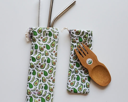 Reusable bamboo spork and stainless steel straw pouch set. The pouches are both white with tiny cartoon avocados printed all over. Some are whole avocados, others are cut in half, and some are slices. Pits are scattered around as well.