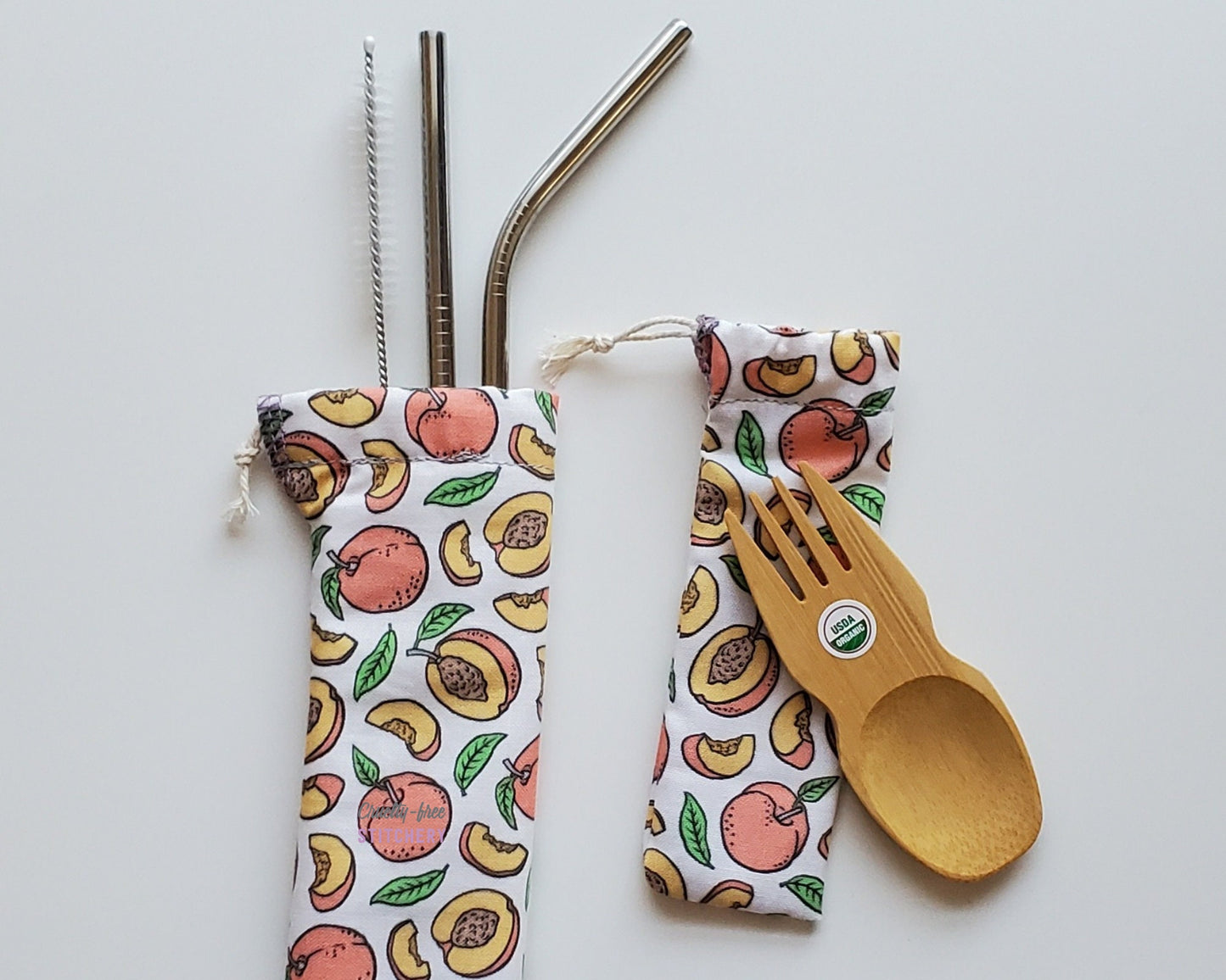 Reusable bamboo spork and stainless steel straw pouch set. The pouches are both white with small peaches all over. Some peaches are whole, others are cut in half or in slices, and some leaves are scattered around as well.
