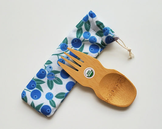 Reusable bamboo spork with pouch. The pouch is a white fabric with blueberry vines printed on. The pouch is sitting diagonally with the spork partially on top pointing the other way. The spork is small, a double ended fork and spoon.