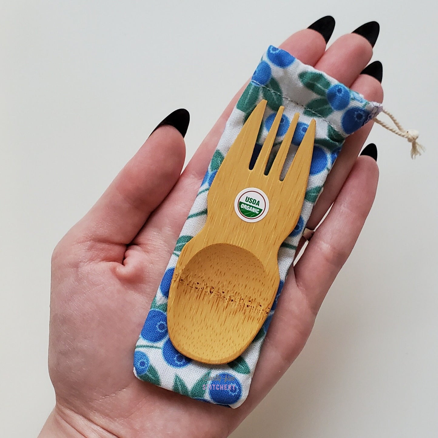 Reusable bamboo spork with pouch. Blueberry print fabric pouch with bamboo spork on top, laid on a hand for size reference. The spork is slightly longer than the fingers.