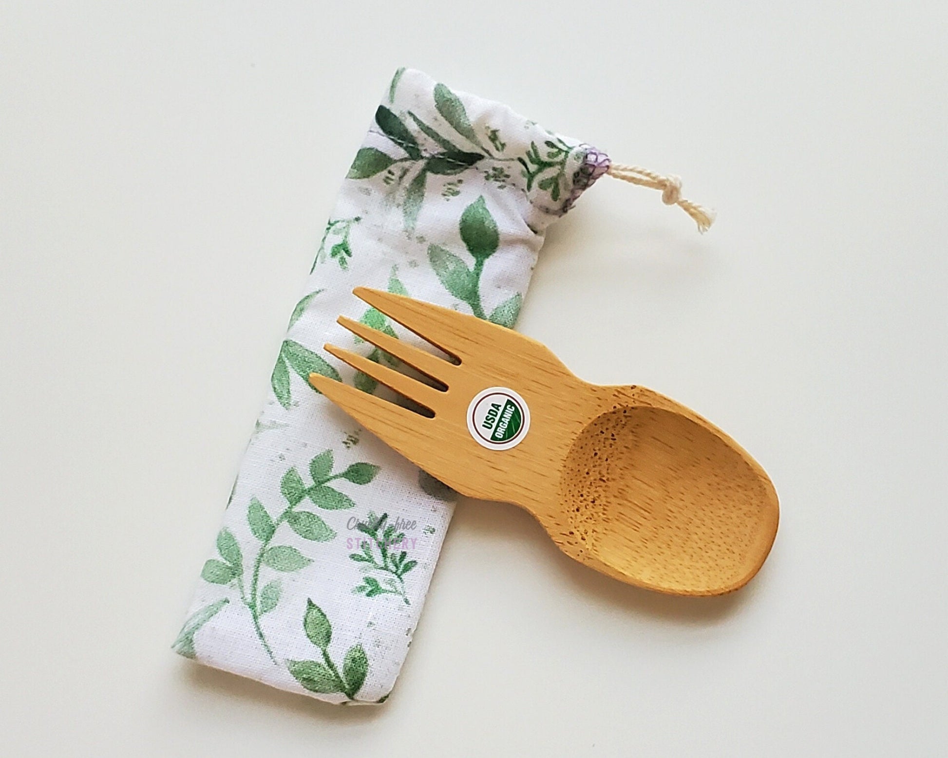 Reusable bamboo spork with pouch. The pouch is a white fabric with green eucalyptus leaves and vines. The pouch is sitting diagonally with the spork partially on top pointing the other way. The spork is small, a double ended fork and spoon.